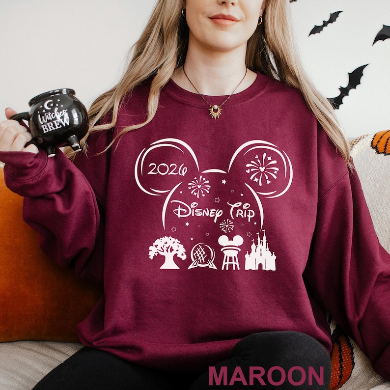 A woman wearing a maroon sweater with a Disney-themed design, including a mug and a chair, is sitting on a couch.