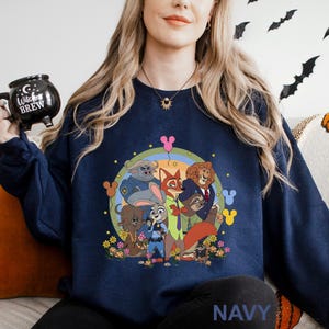 A woman wearing a navy blue sweatshirt with a colorful graphic of cartoon characters from the Disney movie "Jurassic Park" on it. She is holding a black mug with the words "Caffeine" and "Brew" printed on it.