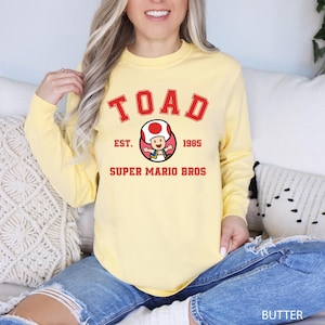 A woman wearing a yellow sweatshirt with a cartoon character and text printed on it, sitting on a couch.