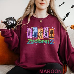 A woman wearing a burgundy sweatshirt with a colorful graphic design featuring characters from the movie "Zootopia 2". She is holding a black mug with the text "Cocktail BRW" on it.