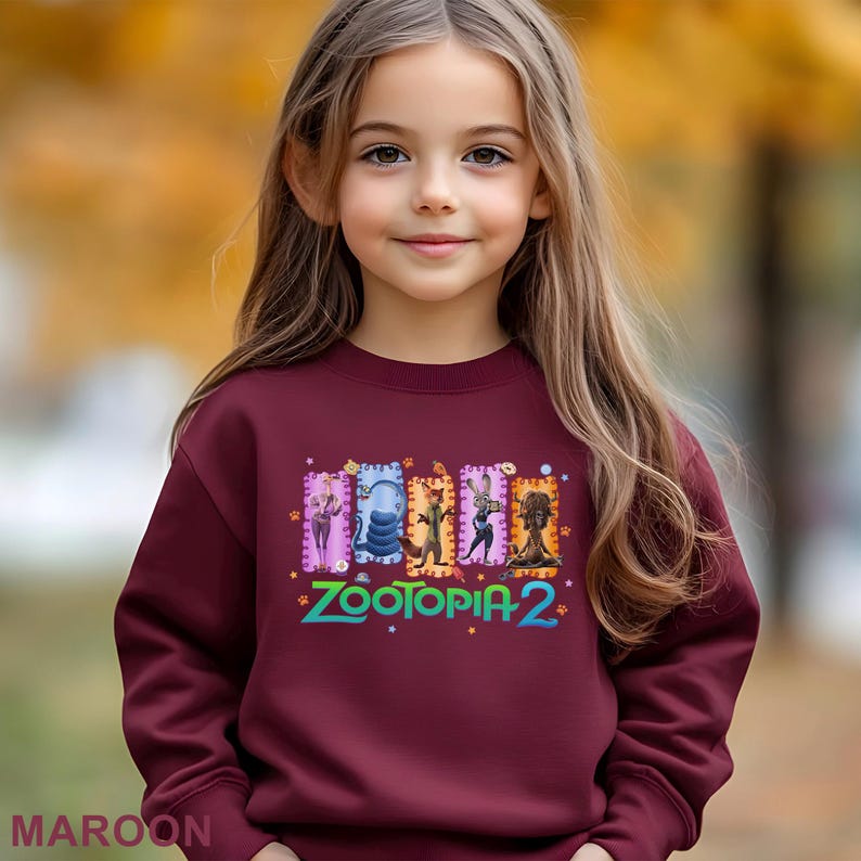 A young girl with long blonde hair is wearing a maroon sweater with a colorful graphic of four cartoon animal characters from the movie "Zootopia 2".