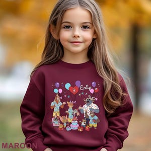 A young girl wearing a maroon sweater with a birthday party design, including animals and balloons.