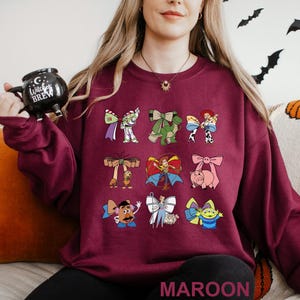 A woman wearing a maroon sweater with various cartoon characters printed on it, holding a black mug.