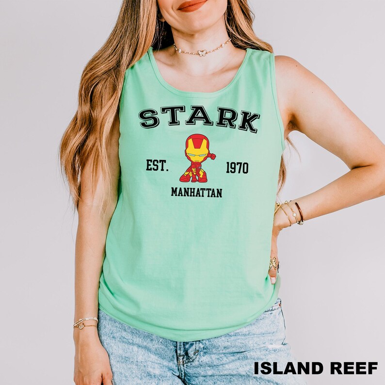 A woman wearing a green tank top with a graphic of a character from the Marvel Cinematic Universe, specifically Iron Man, and text that says "STARK EST. 1970 MANHATTAN ISLAND REEF".