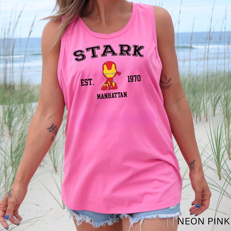 A woman wearing a pink tank top with a graphic of a character from the Marvel Cinematic Universe, specifically Iron Man, standing on a beach.