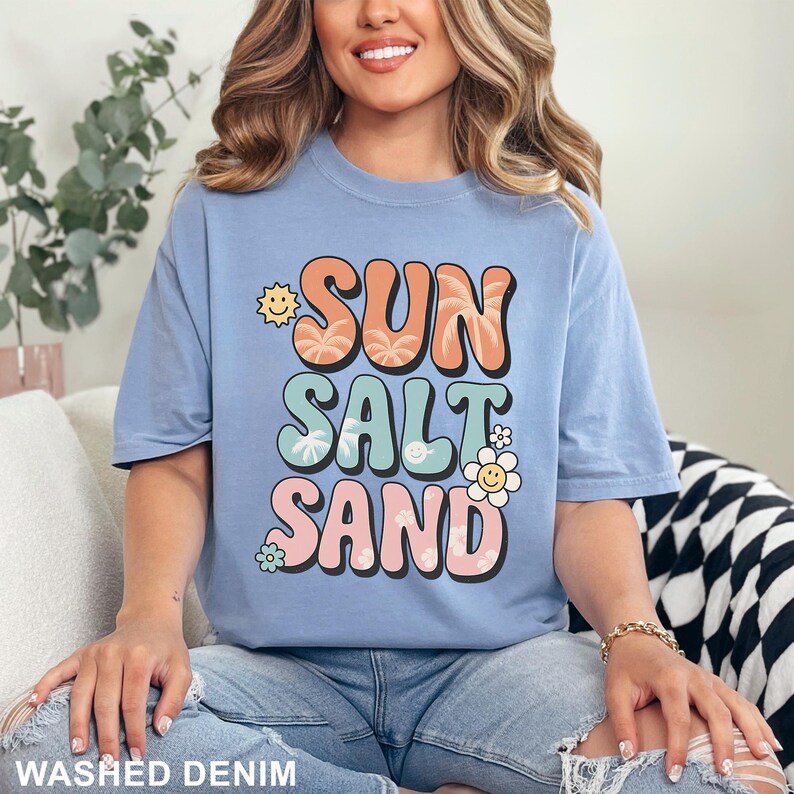 A woman wearing a blue t-shirt with the text "SUN SALT SAND" printed on it, sitting on a couch.