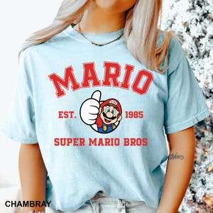 A person wearing a light blue t-shirt with a graphic of Mario and text that says "MARIO EST. 1985 SUPER MARIO BROS".