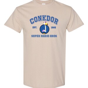 The image is a beige t-shirt with a graphic design featuring a blue circle with a yellow bird inside, and text above and below the circle.