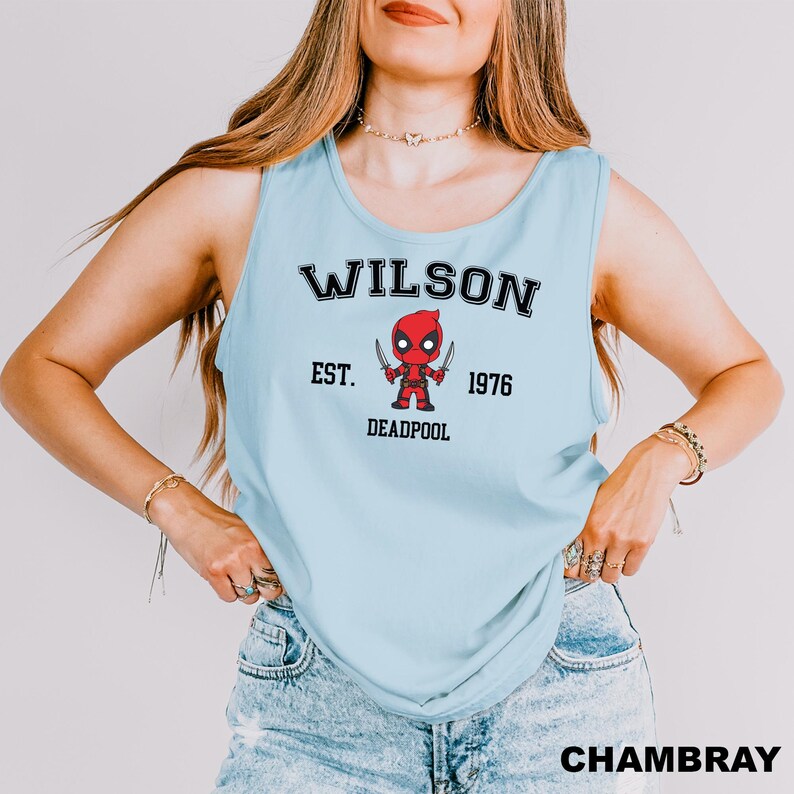 A woman wearing a light blue tank top with a graphic of a character from the movie Deadpool and text that says "Wilson Est. 1976 Deadpool".