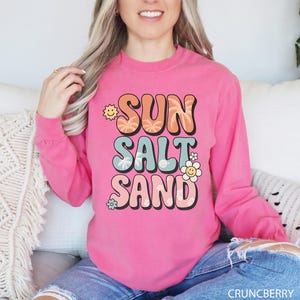 A woman wearing a pink long-sleeved shirt with the text "SUN SALT SAND" printed on it.