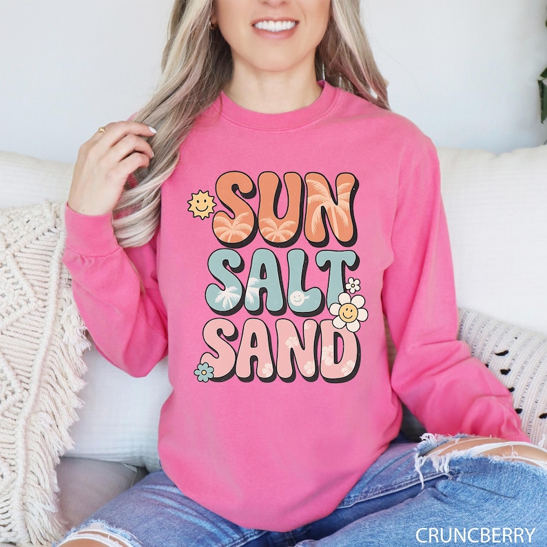 A woman wearing a pink long-sleeved shirt with the text "SUN SALT SAND" printed on it.
