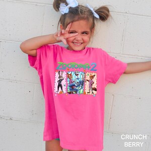 A young girl wearing a pink t-shirt with a cartoon character design is smiling and making a peace sign with her fingers.