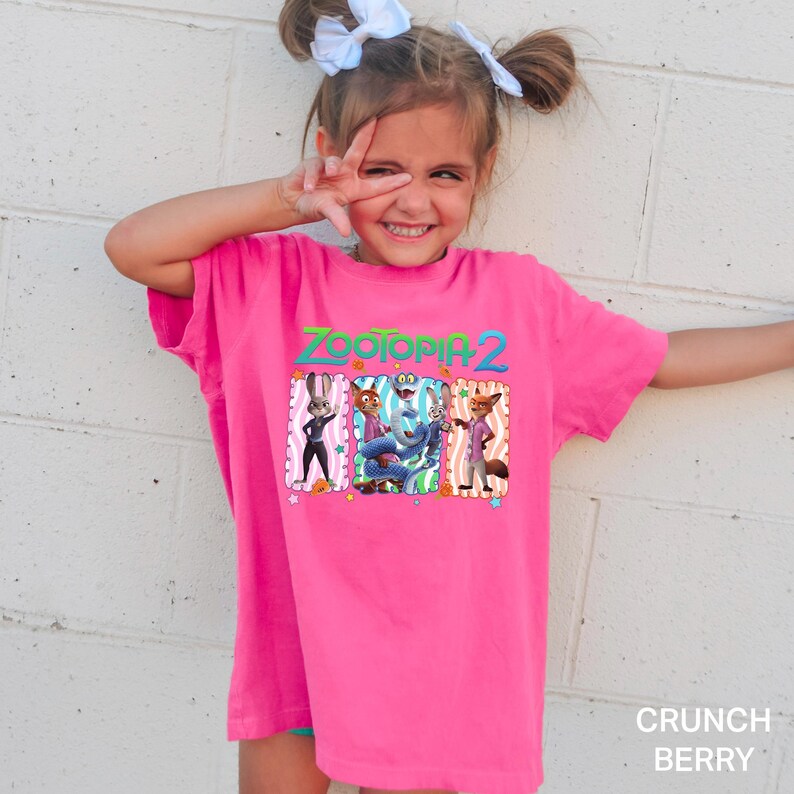A young girl wearing a pink t-shirt with a cartoon character design is smiling and making a peace sign with her fingers.