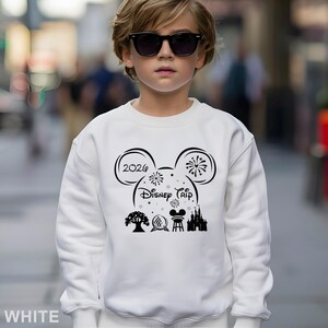 A young boy wearing a white sweater with a Disney-themed design and text, standing on a city street.