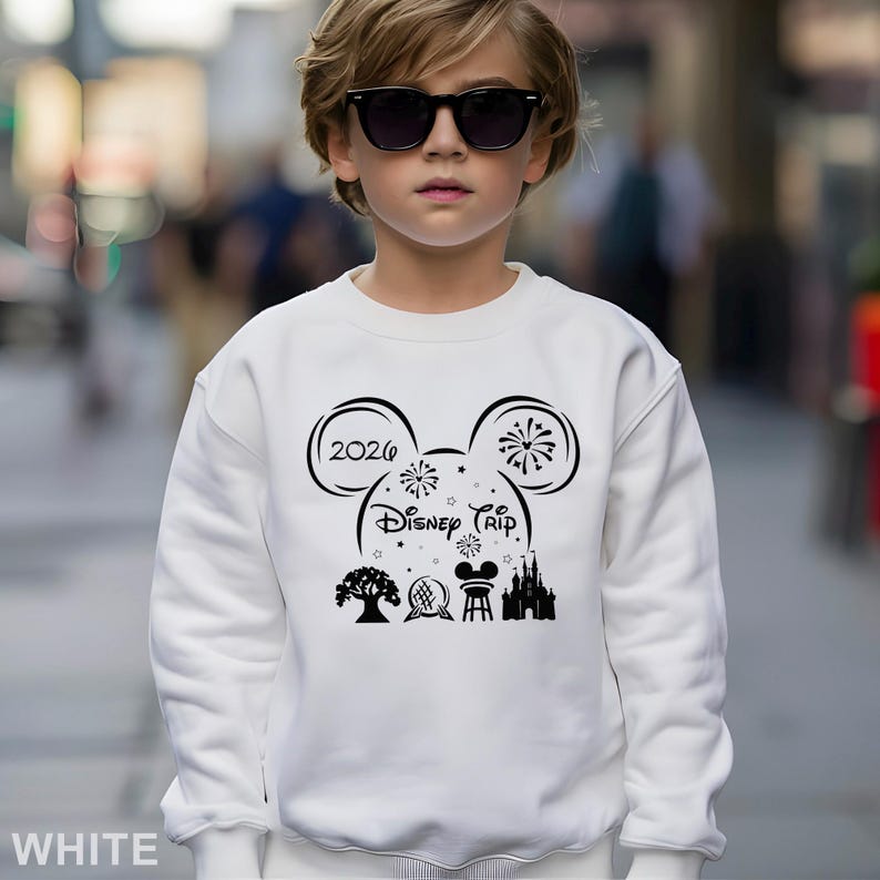 A young boy wearing a white sweater with a Disney-themed design and text, standing on a city street.