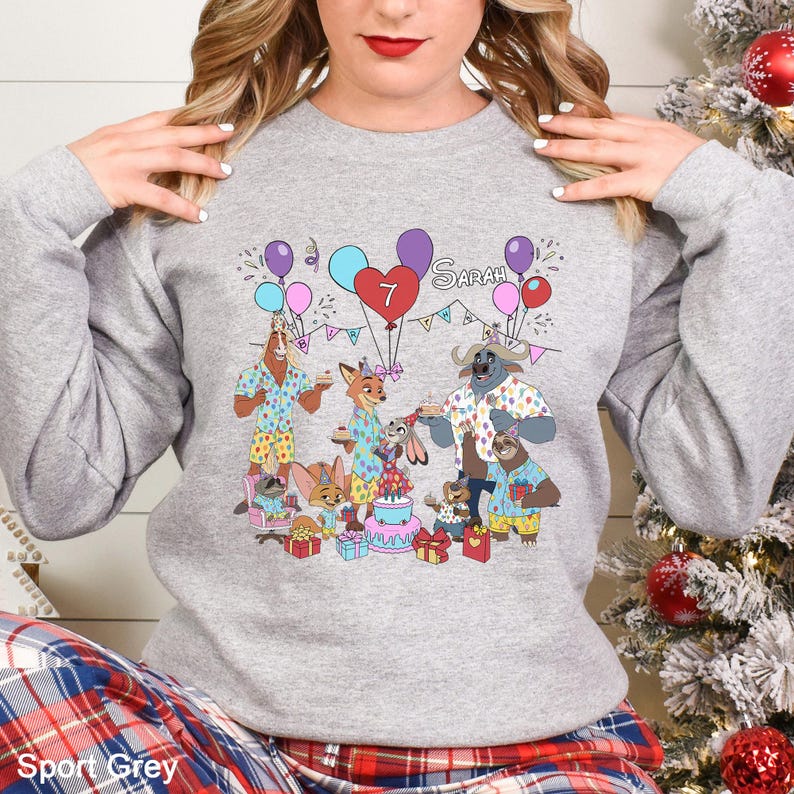 A woman wearing a gray sweatshirt with a cartoon illustration of a birthday party scene featuring various animals and a cake.