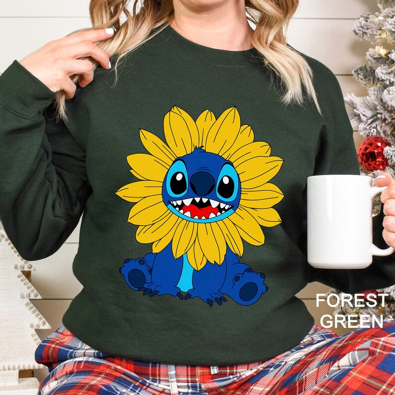 A woman wearing a green sweatshirt with a cartoon character of a blue and yellow flower on it, holding a white mug.