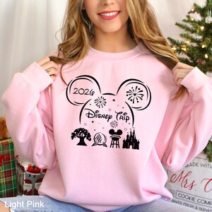 A woman wearing a pink sweatshirt with a Disney-themed design, including a Mickey Mouse ear, fireworks, and a castle.