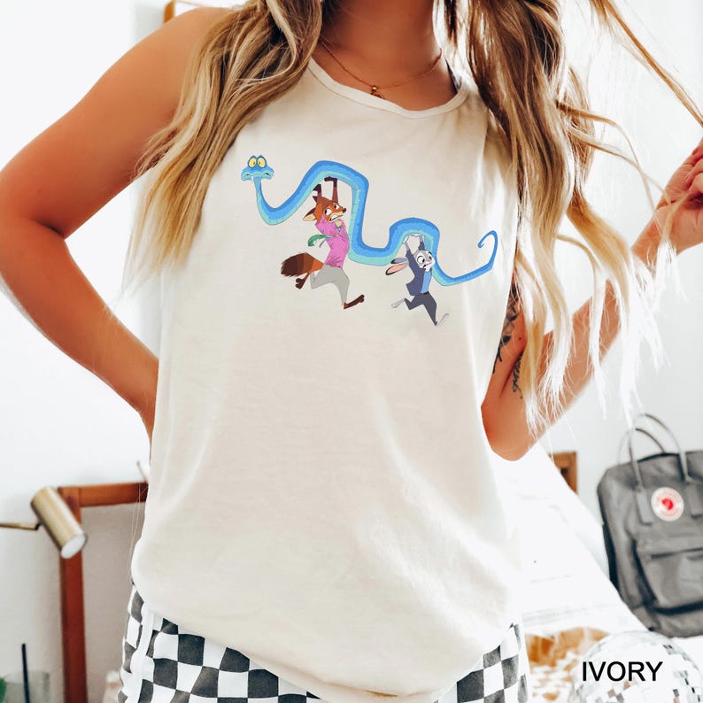 A woman wearing a white tank top with a cartoon character design, standing in a room with a bed and a backpack visible in the background.