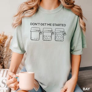 May include: A light green t-shirt with the text "DON'T GET ME STARTED" above a graphic of three jars. The jars have varying contents and small tags. The shirt is a relaxed fit.