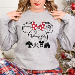 A woman wearing a gray sweatshirt with a graphic design featuring a red bow, fireworks, and Disney-themed icons.