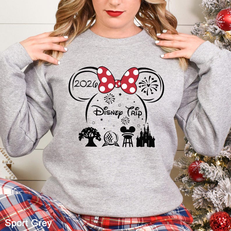 A woman wearing a gray sweatshirt with a graphic design featuring a red bow, fireworks, and Disney-themed icons.