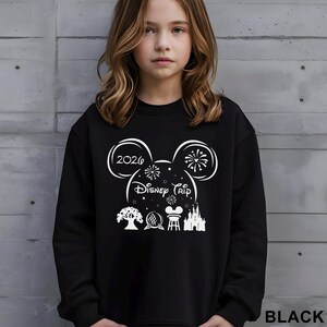 A young girl wearing a black sweater with a graphic design featuring Disney-themed elements such as Mickey Mouse, fireworks, and a castle.