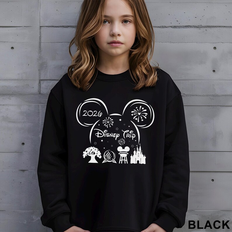 A young girl wearing a black sweater with a graphic design featuring Disney-themed elements such as Mickey Mouse, fireworks, and a castle.