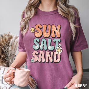 A woman wearing a purple t-shirt with the text "SUN SALT SAND" printed on it, holding a pink coffee mug.