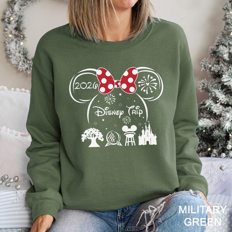 A woman wearing a green sweatshirt with a graphic design featuring a red bow, a tree, a castle, and fireworks.