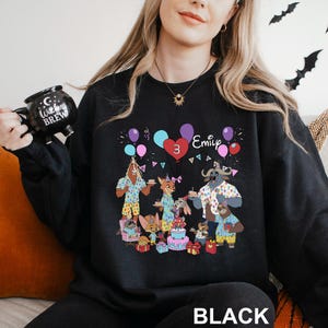 A woman wearing a black sweatshirt with a colorful cartoon character design and the text "Emilie 3" printed on it. She is holding a black mug with the text "Witches Brew" printed on it.