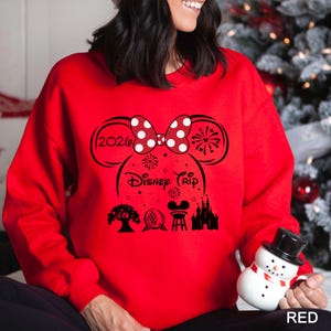 A woman wearing a red sweater with a Disney-themed design, including a Minnie Mouse, fireworks, and a castle, is holding a snowman and a mug.