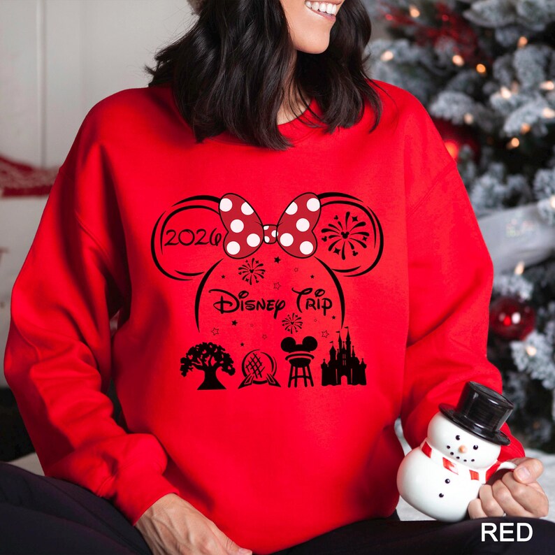 A woman wearing a red sweater with a Disney-themed design, including a Minnie Mouse, fireworks, and a castle, is holding a snowman and a mug.