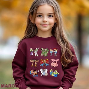 A young girl with long blonde hair wearing a burgundy sweater with various cartoon characters printed on it.