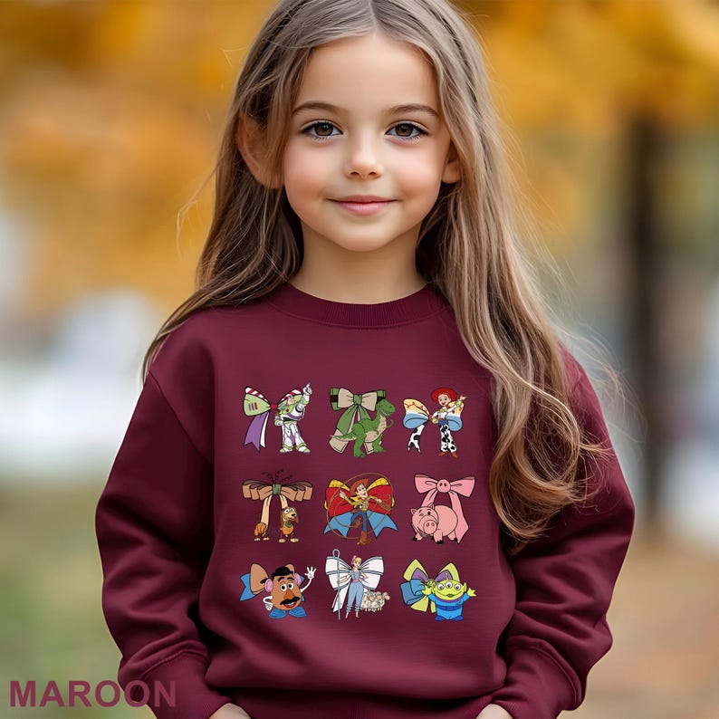 A young girl with long blonde hair wearing a burgundy sweater with various cartoon characters printed on it.