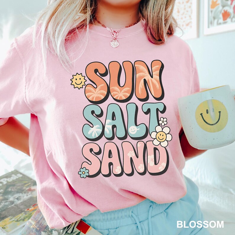 A person wearing a pink t-shirt with the text "SUN SALT SAND" printed on it, holding a coffee mug with a smiley face.