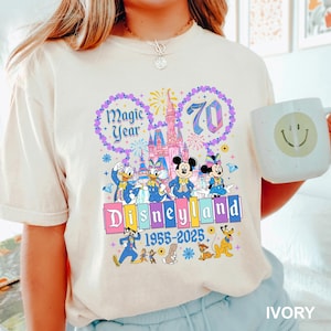 A person wearing a white t-shirt with a colorful graphic design featuring Disney characters and the text "Magic Year" and "Disneyland 1955-2025".