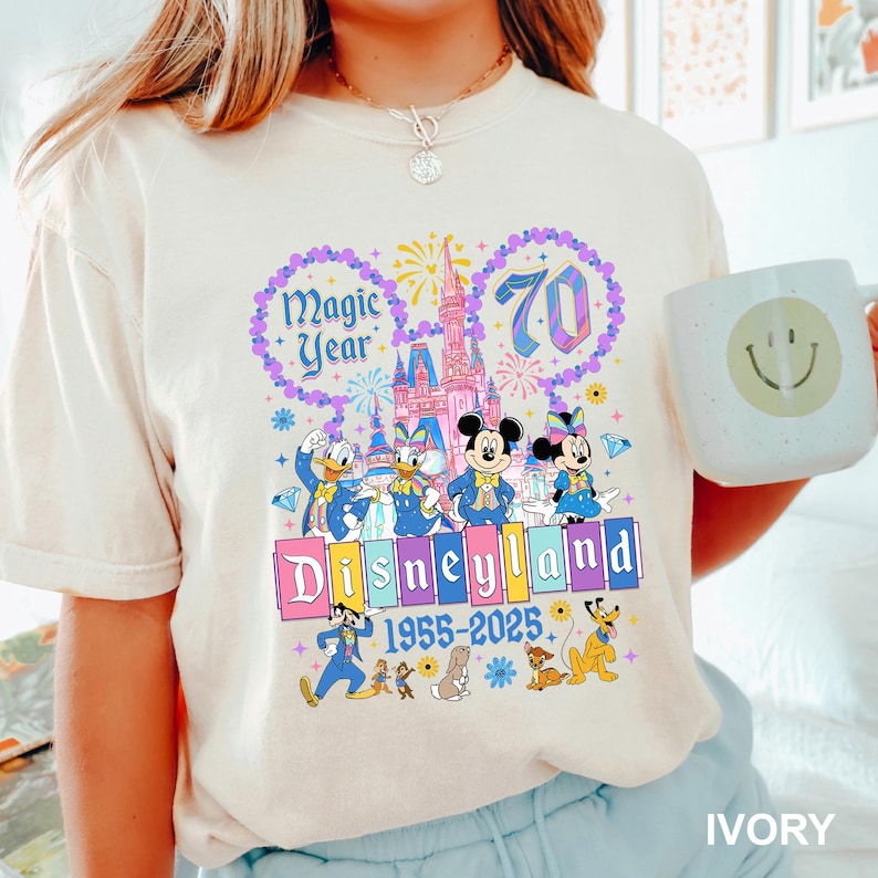 A person wearing a white t-shirt with a colorful graphic design featuring Disney characters and the text "Magic Year" and "Disneyland 1955-2025".