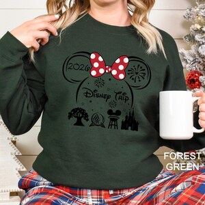 A woman wearing a green sweater with a Disney-themed design, including a red bow, fireworks, and various Disney landmarks, is holding a white mug.