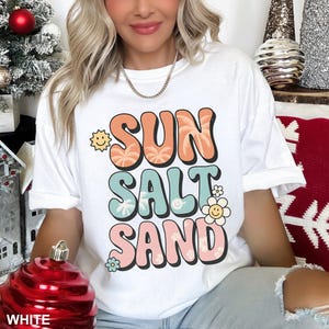 A woman wearing a white t-shirt with the text "SUN SALT SAND" printed on it, sitting in front of a Christmas tree.