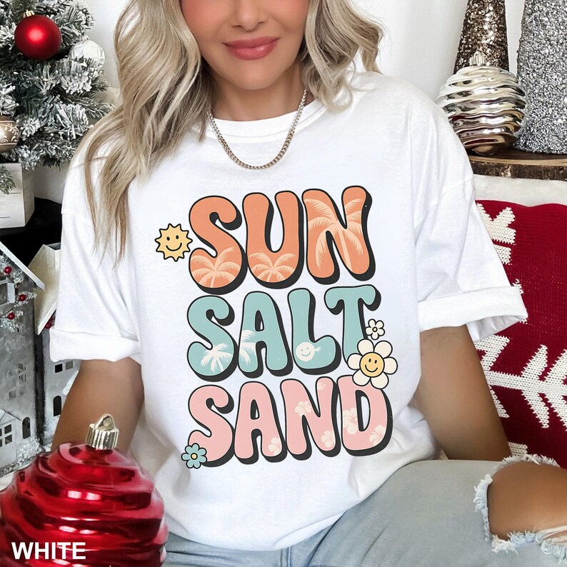 A woman wearing a white t-shirt with the text "SUN SALT SAND" printed on it, sitting in front of a Christmas tree.