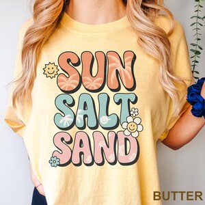 A woman wearing a yellow t-shirt with the text "SUN SALT SAND" printed on it.