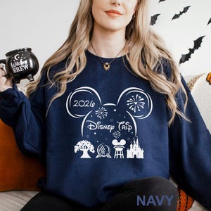 A woman wearing a navy blue sweatshirt with a white design featuring Disney-themed graphics and text, including a Mickey Mouse ear, fireworks, and a castle.