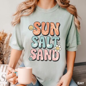 A woman wearing a t-shirt with the text "SUN SALT SAND" printed on it, holding a coffee mug.
