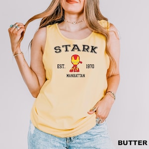 A woman wearing a yellow tank top with a graphic of a character from the Marvel Cinematic Universe, specifically Iron Man, and text that says "STARK EST. 1970 MANHATTAN".