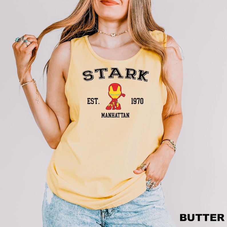 A woman wearing a yellow tank top with a graphic of a character from the Marvel Cinematic Universe, specifically Iron Man, and text that says "STARK EST. 1970 MANHATTAN".