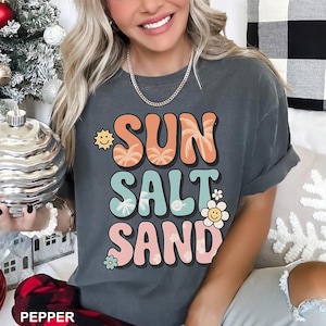 A woman wearing a gray t-shirt with the text "SUN SALT SAND" printed on it, smiling and holding a small object.