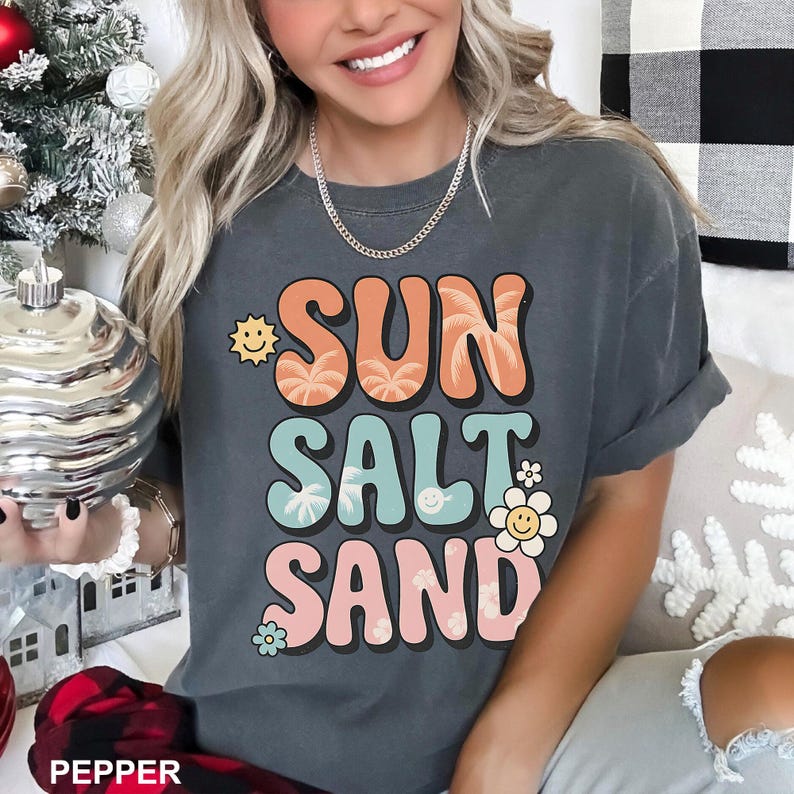 A woman wearing a gray t-shirt with the text "SUN SALT SAND" printed on it, smiling and holding a small object.