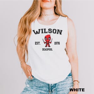 A woman wearing a white tank top with a graphic of a character from the Marvel Cinematic Universe, specifically Deadpool.