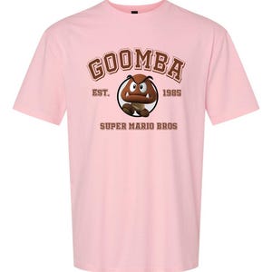 A pink t-shirt with a graphic of Goomba, a character from the Super Mario Bros. video game franchise, and text that reads "GOOMBA EST. 1985 SUPER MARIO BROS".